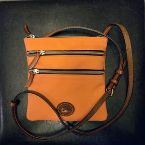 May include: An orange crossbody bag with two horizontal zippered front pockets and a long brown leather adjustable strap. The bag features a small oval leather patch at the bottom center with the embossed text Dooney & Bourke and a duck logo.