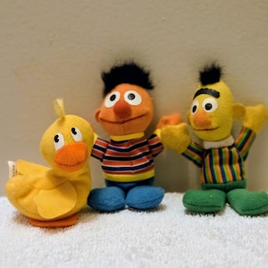 May include: Three Sesame Street plush toys: a yellow duck, an orange-skinned plush doll, and a yellow plush doll. The duck has an orange beak and a small yellow tuft. The orange doll wears a striped shirt and blue trousers. The yellow doll wears a striped shirt and green trousers.