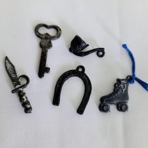 Pode incluir: Uma coleção de pequenos amuletos de metal escuro. Inclui uma chave, uma ferradura, um patim, uma faca e um cachimbo. O patim tem uma corda azul presa. Os amuletos estão sobre uma superfície branca.
