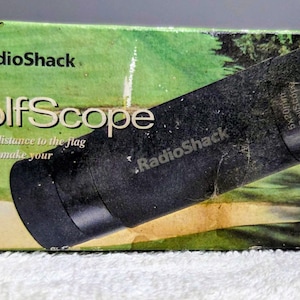 Pode incluir: Uma caixa RadioShack GolfScope com um telêmetro de golfe preto. A caixa verde exibe o logotipo da RadioShack, a palavra "GolfScope" e o texto "Shows the distance to the flag so you can make your best shot!"