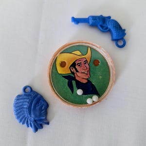 Op de afbeelding: Een verzameling vintage speelgoedbedels en een spel. Inclusief een blauw plastic speelgoedpistool, een blauwe Native American profielbedel en een rond spel met een cowboyafbeelding en witte ballen.