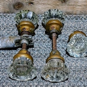 May include: Antique glass doorknobs with brass hardware. The doorknobs have a faceted design and are clear. One doorknob is attached to a metal plate. The hardware shows signs of age and wear.