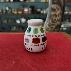 Könnte beinhalten: Kleine, weiße Keramikvase mit zylindrischer Form. Die Vase zeigt eine Reihe von Farbmustern in Grün, Rot und Lila, mit entsprechenden Nummern und dem Wort "LAZE" auf der Vorderseite. Die Vase steht auf einer roten Oberfläche.