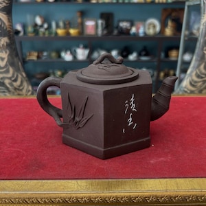 May include: A brown, square-shaped teapot with a lid and handle. The teapot has a textured surface and features an embossed design on one side. The teapot is sitting on a red surface with a gold frame.