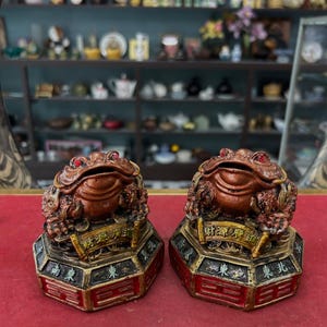 May include: Two ornate, brown frog figurines with red gemstone eyes. Each frog sits atop a gold and red base with Chinese characters. The frogs are displayed on a red surface, with a blurred background of shelves filled with various items.