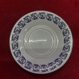 May include: White saucer with a blue repeating pattern around the rim. The pattern consists of a stylized monogram. The saucer has a gold rim and a recessed center for a cup.