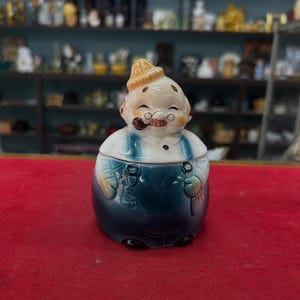 Könnte beinhalten: Ein Vintage-Keksbehälter aus Keramik in Form eines lächelnden alten Mannes mit einer Pfeife. Der Behälter hat einen weißen Kopf mit einem beigen Hut, runden Gläsern und einem schwarzen Schnurrbart. Der Körper ist eine blaue Latzhose über einem weißen Hemd.