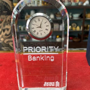 Pode incluir: Um relógio de mesa de vidro transparente com movimento a quartzo. O mostrador é branco com algarismos romanos e ponteiros pretos. As palavras "PRIORITY Banking" estão impressas no vidro. O logotipo Standard Chartered está na parte inferior.