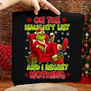 May include: Black pillow with a Christmas-themed graphic. The design features a green character in a Santa hat, red suit, and holding a glass of wine, with the text "ON THE NAUGHTY LIST AND I REGRET NOTHING."