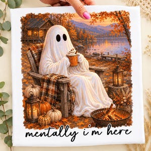 Op de afbeelding: Een wit t-shirt met een afbeelding van een geest die in een houten stoel bij een meer zit en een mok vasthoudt. De scène bevat een hut, pompoenen, lantaarns en een vuurkorf. De tekst "mentally i'm here" staat in het zwart onderaan.