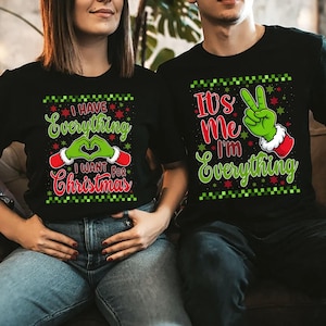 Puede incluir: Camisetas negras con gráficos navideños. Una camiseta dice "I Have Everything I Want For Christmas" con manos verdes formando un corazón. La otra dice "It's Me I'm Everything" con una mano verde haciendo el signo de la paz.