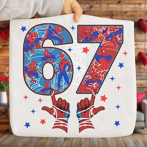 Może przedstawiać: Biała torba z numerem 67, ozdobiona postaciami Spider-Mana i pajęczynami. Numer otoczony jest czerwonymi i niebieskimi gwiazdami. Dwie dłonie Spider-Mana wskazują w górę.