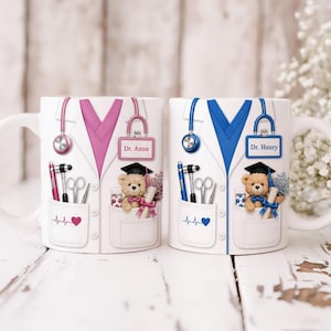 Puede incluir: Dos tazas de cerámica blanca con temática de médico. Una taza presenta un diseño de bata rosa, la otra un diseño de bata azul. Cada taza tiene un estetoscopio, un bolsillo con herramientas médicas y un osito de peluche con birrete. Las tazas están etiquetadas como "Dr. Anna" y "Dr. Henry".
