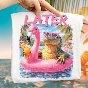 May include: White tote bag featuring a whimsical illustration of an alligator wearing heart-shaped sunglasses and floating in a pool on a pink flamingo float. The alligator is holding a cocktail. The word "LATER" is printed above the image.