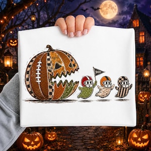 May include: A white t-shirt featuring a graphic of a large, textured pumpkin designed like a football, with its mouth open as if chasing three small ghosts wearing football helmets. One ghost holds a small flag with the text BOO printed on it.