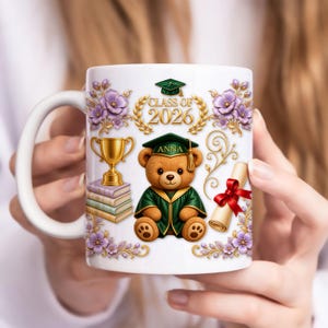 May include: White ceramic mug with a graduation theme. The design includes a teddy bear in a graduation gown, the text "Class of 2026", a diploma, a trophy, and floral accents. The mug is held by a person.