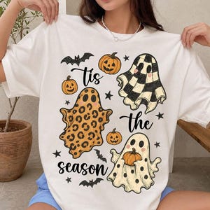 Può includere: Una t-shirt bianca a maniche corte con una stampa grafica che riporta il testo 'Tis the season'. Il design include tre fantasmi con motivi diversi, zucche arancioni, pipistrelli neri e piccole stelle nere sparse sul davanti.