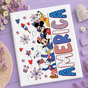 May include: A white rectangular print featuring the word AMERICA in large, stylized letters filled with red, white, and blue patterns. The design includes illustrations of cartoon characters, fireworks, small flags, and balloons in a patriotic theme.