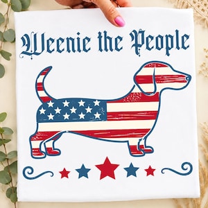 Può includere: Una t-shirt bianca con la grafica di un cane bassotto riempito con un motivo a bandiera americana rossa, bianca e blu. Sopra il cane, il testo Weenie the People in un carattere gotico blu scuro. Sotto il cane ci sono cinque stelle e ghirigori.