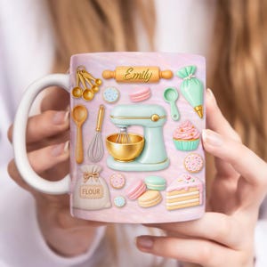 May include: A white ceramic mug featuring a pink background with baking-themed illustrations. The design includes a stand mixer, rolling pin with the name "Emily", measuring spoons, and various baked goods like cookies and a cupcake.