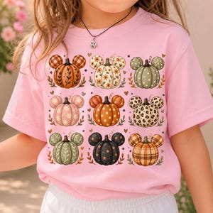 Puede incluir: Una camiseta de manga corta de color rosa claro con una cuadrícula de nueve ilustraciones de calabazas, cada una con forma de orejas de ratón. Las calabazas muestran varios patrones como cuadros, flores, lunares y estampado de leopardo.