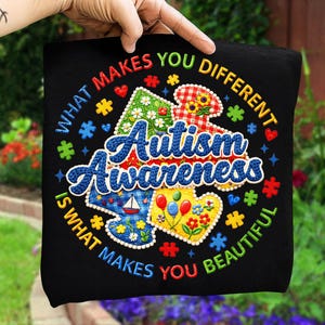 May include: Black t-shirt with embroidered text and colorful puzzle piece designs. The shirt reads "WHAT MAKES YOU DIFFERENT IS WHAT MAKES YOU BEAUTIFUL" and "Autism Awareness". The design includes hearts, flowers, and a sailboat.