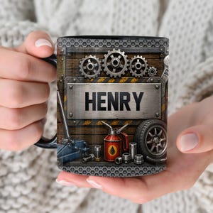 Puede incluir: Taza de café negra con un diseño de temática mecánica. La taza presenta el nombre "HENRY" en una placa de metal, rodeada de engranajes, herramientas y un neumático. El fondo tiene una textura de madera con rayas amarillas y negras.