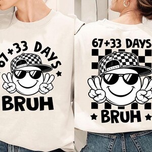 Peut inclure: Sweat-shirt blanc cassé avec un motif d'un visage souriant portant des lunettes de soleil et une casquette à carreaux noir et blanc, faisant le signe de la paix. Le texte "67+33 DAYS BRUH" est imprimé en noir.