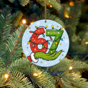 May include: A round Christmas ornament with the numbers 6 and 7 in red and green, decorated with string lights. The ornament has a light blue background with snowflakes and stars, and a green furry design at the bottom.