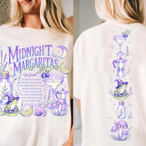 May include: An off-white t-shirt featuring a purple and green graphic print. The front displays the text Midnight Margaritas, a frog in a witch hat, a margarita glass, a bottle, mushrooms, and a snail. The back shows a vertical arrangement of these icons.