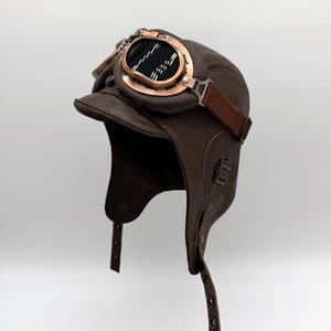 May include: A brown leather aviator hat with attached goggles. The hat has ear flaps, a small peak, and a strap to secure the goggles. The goggles have a copper-coloured frame and dark lenses. The hat is designed in a vintage style.