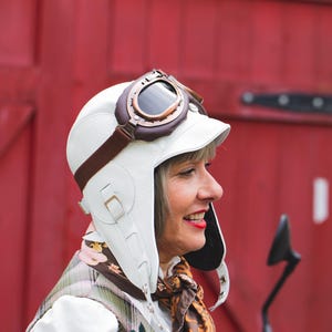 May include: A white leather aviator cap with ear flaps and a short brim, featuring a brown leather strap and buckle detail. The cap is worn with vintage-style brown and copper-toned goggles with dark lenses positioned on the top of the head.