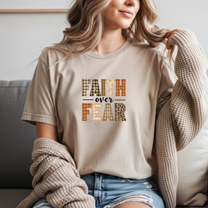 Könnte beinhalten: Beiges T-Shirt mit dem Aufdruck "FAITH over FEAR" auf der Vorderseite. Der Text ist in einer Mischung aus gemusterten Schriftarten gehalten, darunter Karomuster und Leopardenmuster. Das Shirt hat kurze Ärmel und einen Rundhalsausschnitt.