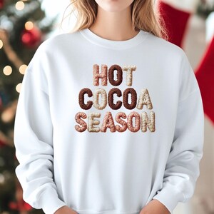 Puede incluir: Sudadera blanca con la frase "HOT COCOA SEASON" en letras texturizadas multicolores. Las letras son en tonos marrón, beige y rosa. Un estilo de ropa casual y cómodo.