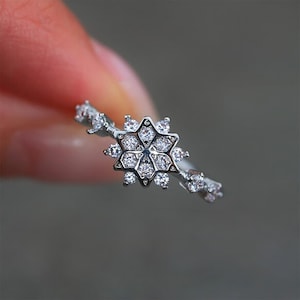 Könnte beinhalten: Ein silberner Ring mit einem Schneeflocken-Design. Der Ring hat ein zartes Band mit kleinen, funkelnden Akzenten. Die zentrale Schneeflocke besteht aus mehreren klaren Edelsteinen und erzeugt einen brillanten, auffälligen Effekt.