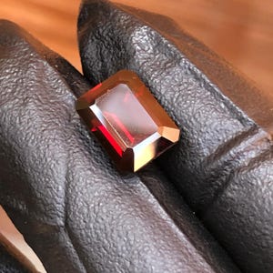 Può includere: Una gemma rettangolare sfaccettata, di un rosso intenso. La gemma ha una superficie lucida ed è tenuta su uno sfondo scuro. Il taglio della pietra rivela molteplici sfaccettature, esaltandone la brillantezza.