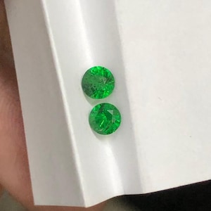 May include: Two round, faceted, bright green gemstones. The gems are displayed on a white surface, with a slight angle, showcasing their brilliant cut and vibrant colour. The gemstones appear to be emeralds or similar precious stones.