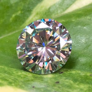 Pode incluir: Uma pedra preciosa redonda e facetada com um brilho multicolorido brilhante. A pedra transparente reflete a luz, exibindo tons de azul, verde e amarelo. A gema está sobre uma folha verde, realçando sua aparência vibrante.
