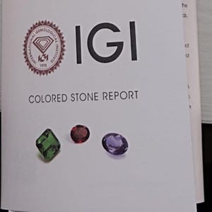 May include: An International Gemological Institute IGI Colored Stone Report document featuring a circular logo, the text IGI, and the words COLORED STONE REPORT. Below the text are three loose gemstones: a square green stone, a round red stone, and an oval purple stone.