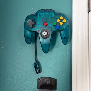 May include: A teal Nintendo 64 controller with a black cable and a black cartridge holder. The controller has a directional pad, red, green, and blue buttons, and a yellow button cluster. The cartridge holder has the letters "N" and "64" on it.