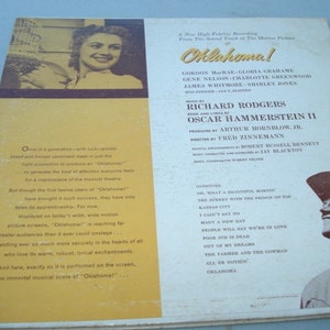 May include: A vintage record album cover for the musical "Oklahoma!" featuring a colour photo of a woman holding a bouquet of flowers. The album cover includes the names of the actors, producers, and composers. The back of the album cover lists the songs included on the record.