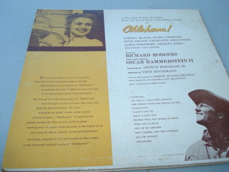 May include: A vintage record album cover for the musical "Oklahoma!" featuring a colour photo of a woman holding a bouquet of flowers. The album cover includes the names of the actors, producers, and composers. The back of the album cover lists the songs included on the record.