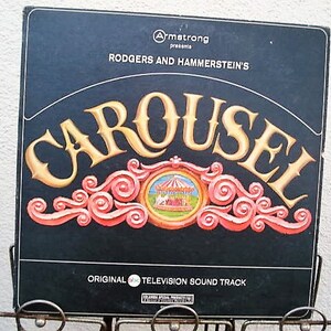 May include: A vintage vinyl record album cover for Rodgers and Hammerstein's "Carousel." The cover is black with gold lettering and a decorative red and gold design. The album is an original television soundtrack.