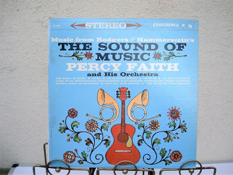 May include: A vintage vinyl record album cover for "The Sound of Music" by Percy Faith and His Orchestra. The album cover is light blue with the title in large white letters. A guitar and horns are featured in the center, with floral accents.