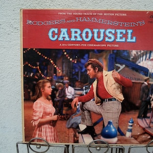 May include: Vintage vinyl record album cover for "Carousel," a Rodgers and Hammerstein musical. The cover features a scene from the 20th Century-Fox Cinemascope picture, with actors in period costumes. The album is displayed in a metal rack.