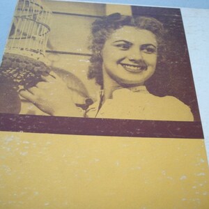 May include: A black and white photograph of a woman with short, curly hair. She is smiling and holding a birdcage. The photo is on a yellow background with a brown border.