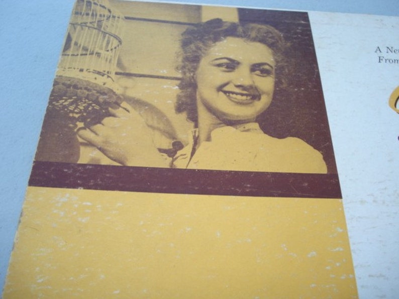 May include: A black and white photograph of a woman with short, curly hair. She is smiling and holding a birdcage. The photo is on a yellow background with a brown border.