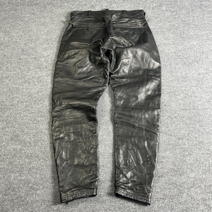 May include: Black leather trousers with a textured finish. The trousers feature a classic five-pocket design, a zip fly, and belt loops. The leather appears worn, adding to the vintage aesthetic. The trousers are laid flat on a grey surface.