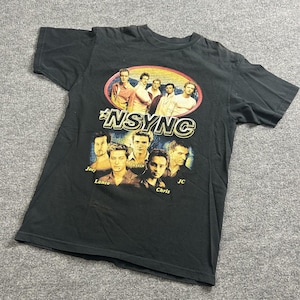 May include: Black t-shirt featuring the boy band *NSYNC. The shirt has a graphic of the band members with the band's logo in a gold and white font. The band members are also featured in an oval design.