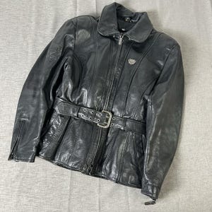 May include: Black leather jacket featuring a belted waist with a silver buckle. The jacket has a zip-up front, a collar, and a small emblem on the chest. The sleeves have zippers at the cuffs. A classic motorcycle-style jacket.